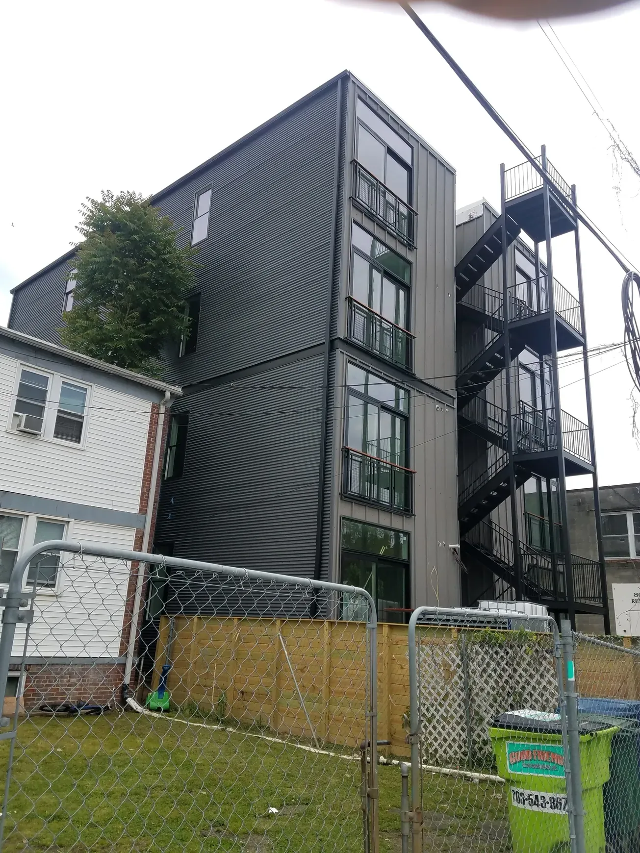 Tall dark-clad building with an exterior metal stair, seen past a fence.